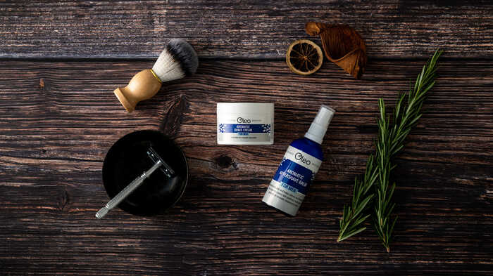 Aromatic shaving cream and aftershave balm by Oleo laid upon a wooden table with shaving equipment and a sprig of rosemary